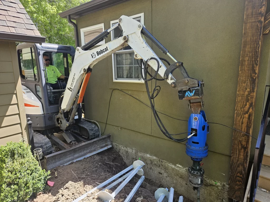 Crew from Utah Screw Pile advancing a helical pier into expansive Utah soils with excavator on residential foundation helical pier installation.